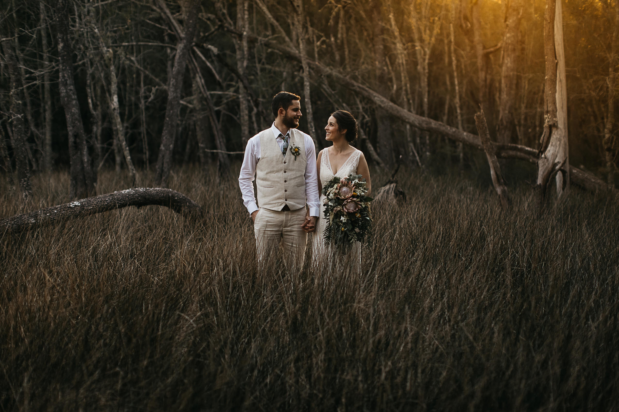 Bride and groom stand amongst trees and grass against the faint glow of the setting sun