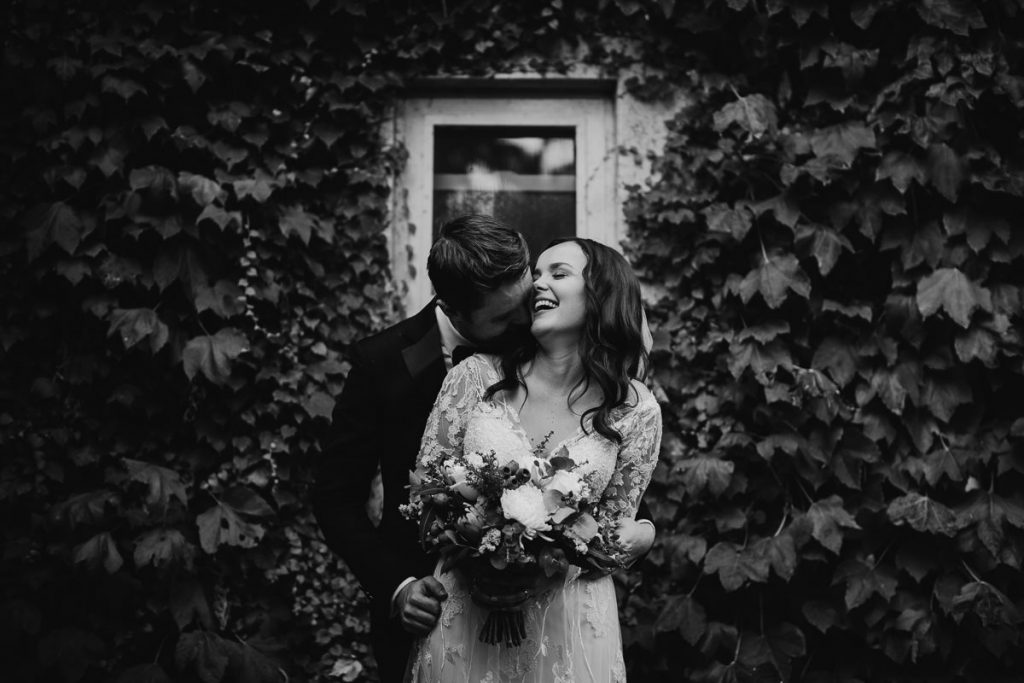 Black and white photo of bridal couple laughing in front of a window and vines at their Bendooley Estate Wedding; photographer