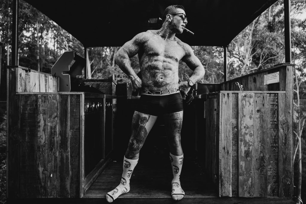 Black and white image of groom in his jocks showing off his tats and his cigar in a BBQ shelter before his wedding at Bundanon on the South Coast
