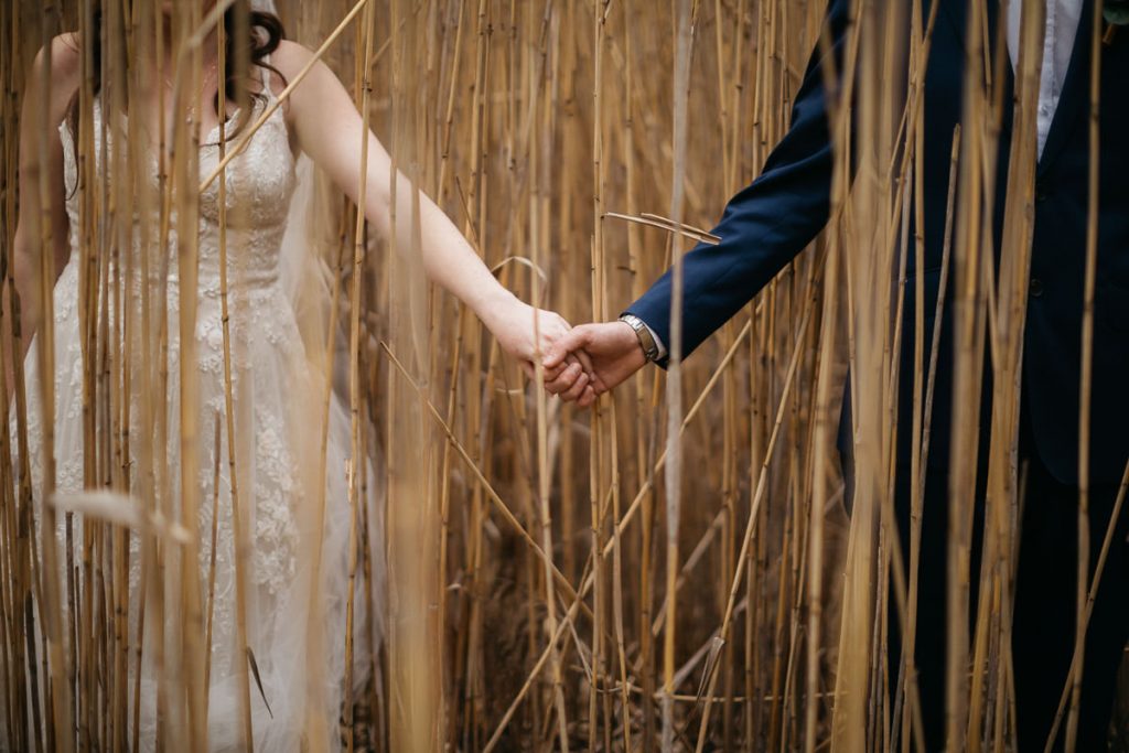 Creative photograph of bride and groom holding hands hidden between the dried wheat at their Butter Factory Wedding on the South Coast