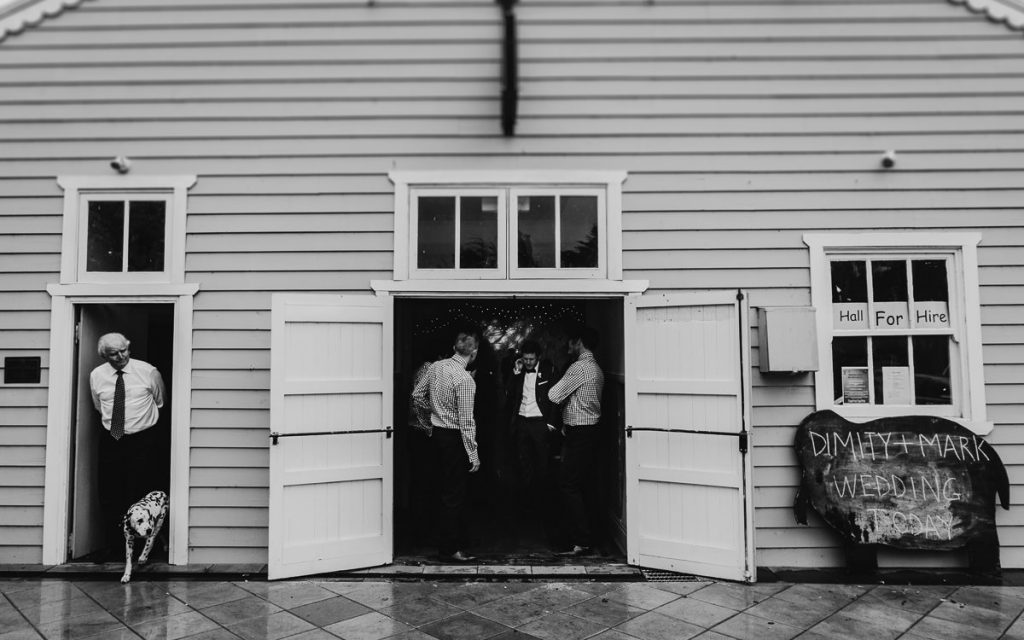 Black and white photograph of a weatherboard hall in Canberra with wedding guests chatting in the open doorways ; Canberra Country Wedding Photographer