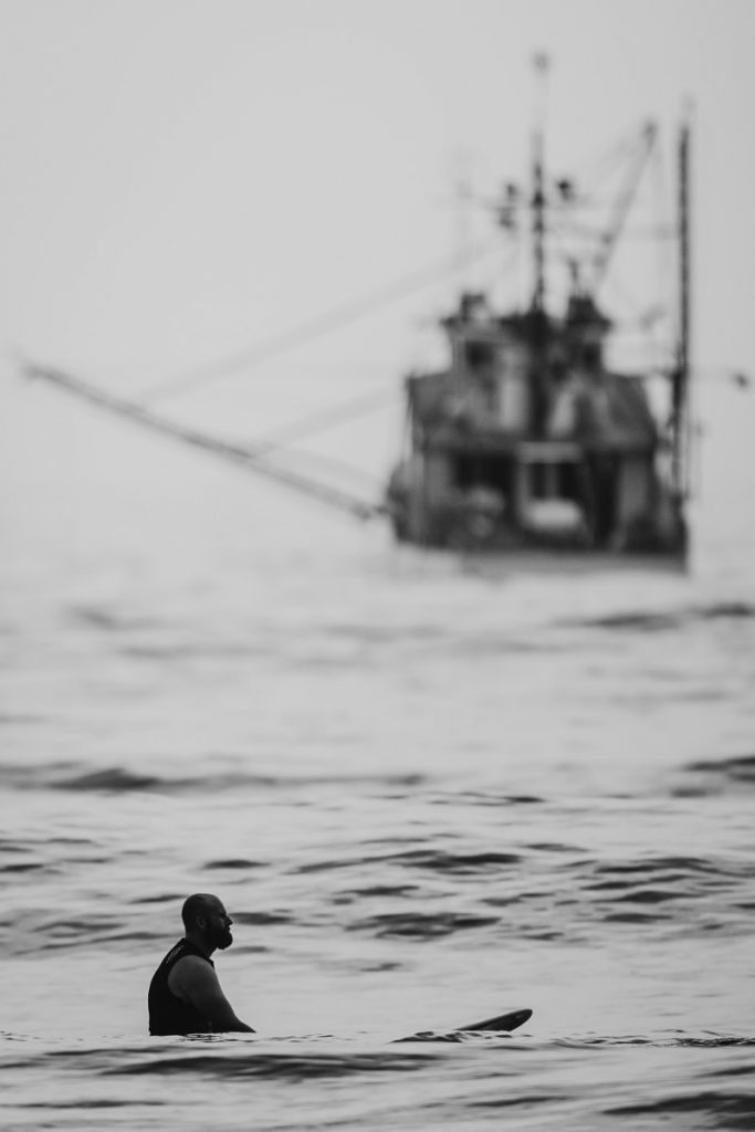 Black and white photograph of grrom sitting thinking on his surfbood with a fishing trawler in the background