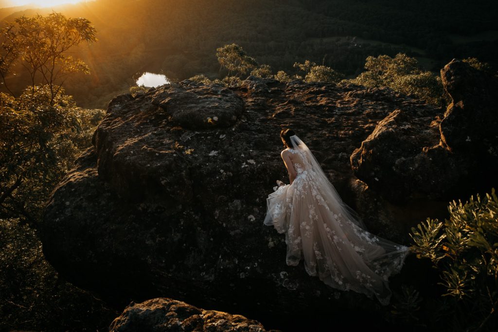 Stunning landscape photograph of bride kneeling, facing the sunset and showing off her wedding gown on the rocks at her Kangaroo Valley Bush retreat Wedding on the South Coast