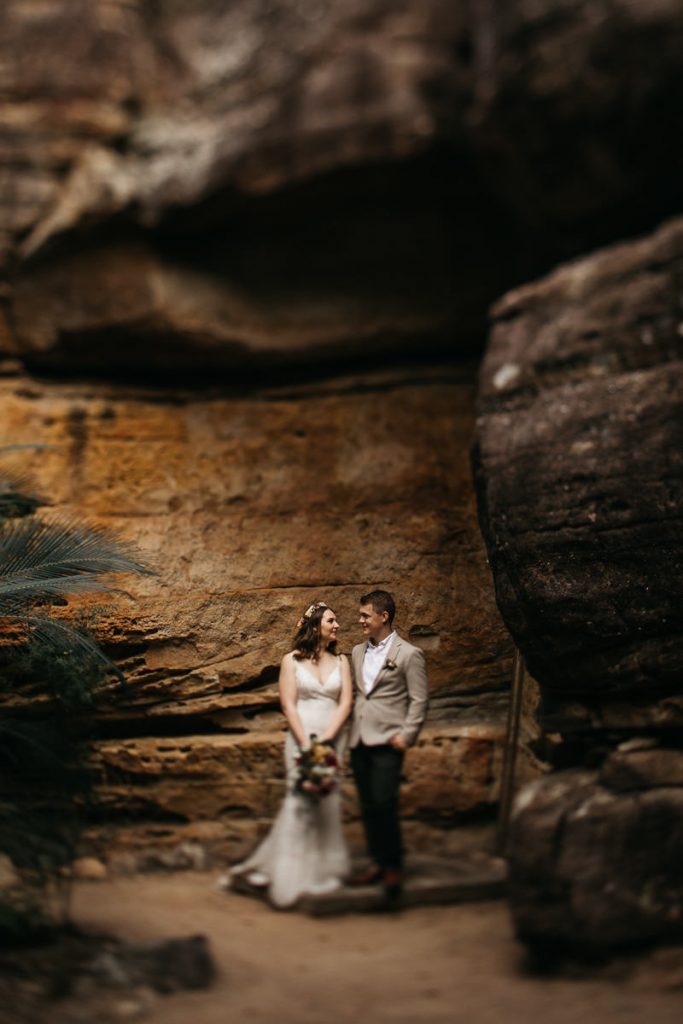bride and groom stand below the rocks at their Kangaroo Valley Wedding; photograph with blurred vignette style finish