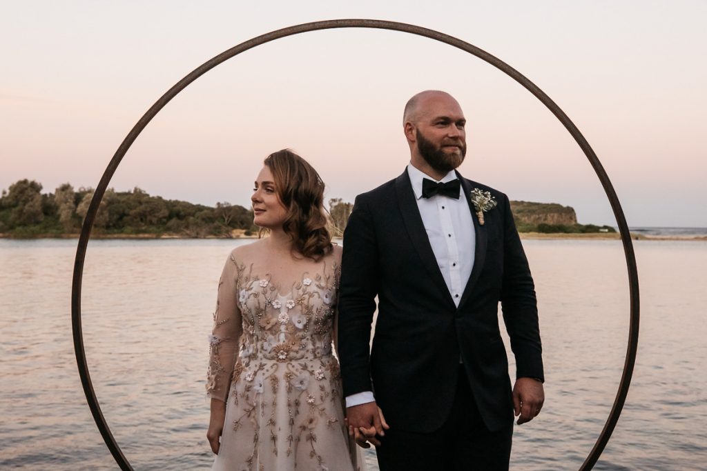 Wedding photography South Coast; Kiama; Bride and groom stand in front of the water with a circular structure framing them at sunset.
