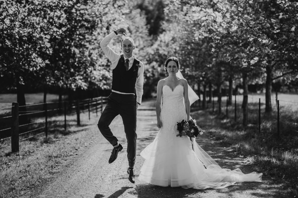 Black and white photo of groom jumping for joy while walking alongside his bride at their Melross Wedding in the Kangaroo Valley - South Coast Wedding Photographer