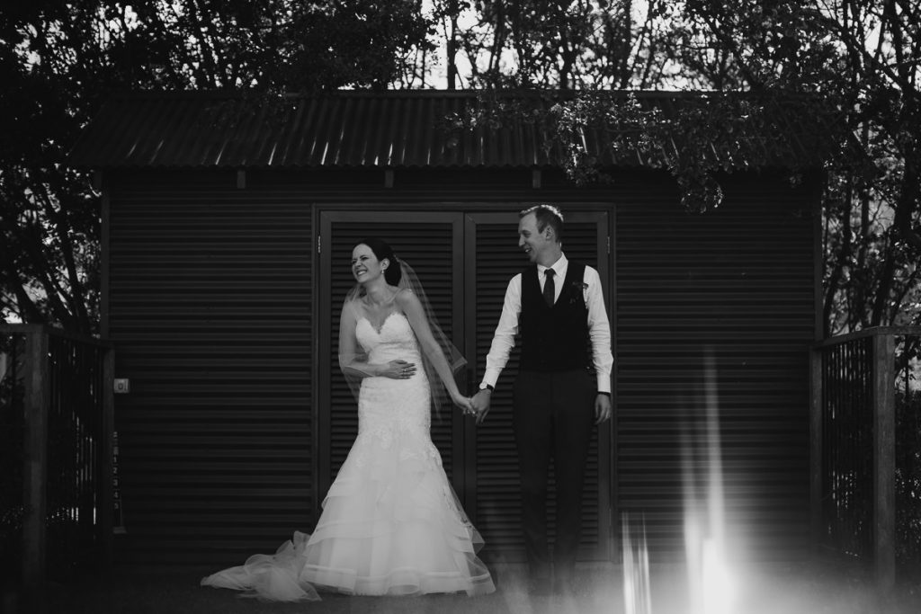 Black and white image of Bride laughing at something the groom said while posing for photos in front of a tin shed; Melross, Kangaroo Valley Wedding Photographer South Coast