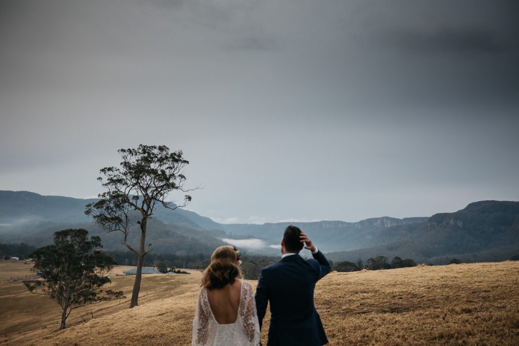 Bride and groom walk across the dry grass towards a stunning view of the Kangaroo Valley at their Melross wedding; South Coast Wedding Photography