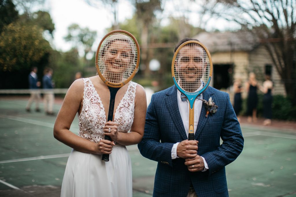 Creative wedding photography of bride and groom posing on a tennis court with old fashioned raquets over their faces in Merribee