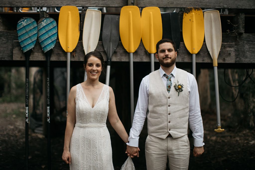 The bridal couple face the photographer smilingly, holding hands, infront of a row of colourful paddles at Paperbark Camp on the South Coast
