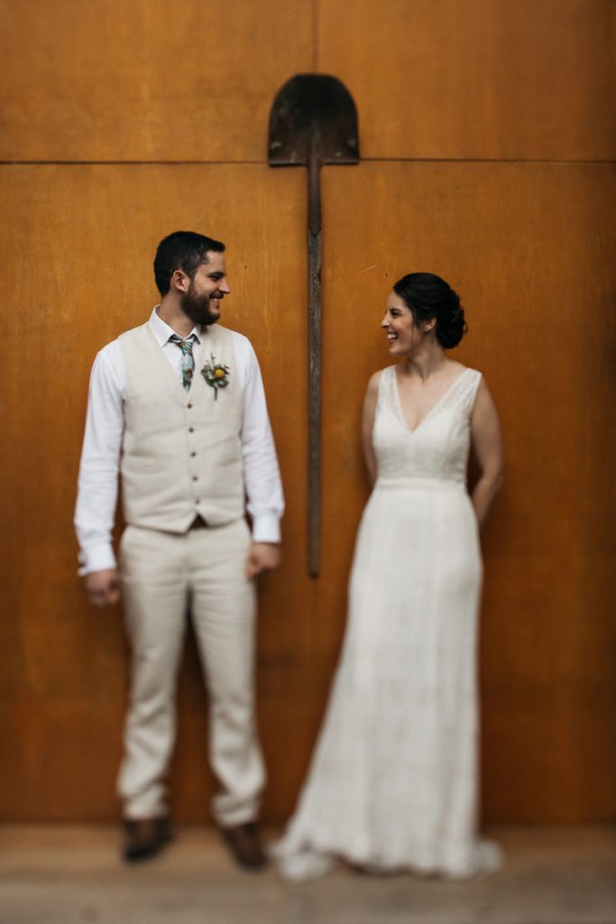 Portrait of bride and groom leaning against a wall with a shovel hanging between them at their Paperbark Camp Wedding; South Coast Wedding Photographer