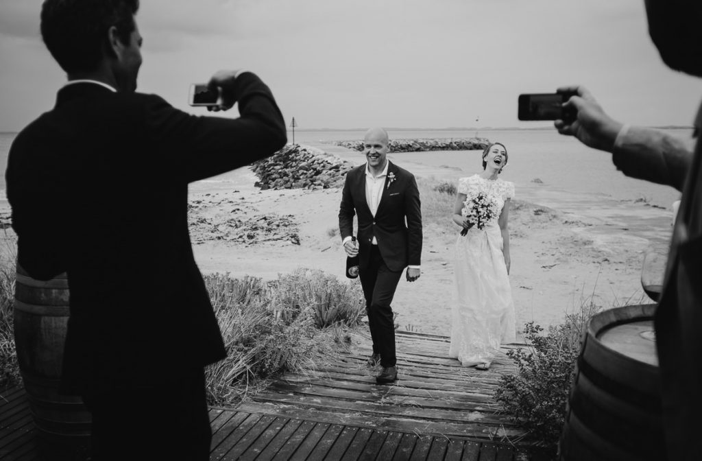 Black and white image of wedding couple walking towards friend photographing them on their mobile phones; South Coast Wedding Photographer
