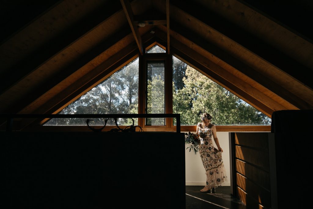 Creative wedding portrait of bride and her floral wedding dress standing in front of a triangular window in the Southern Highlands