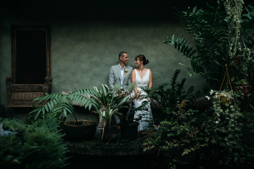 Bride and groom lean against a green wall behind bushes and lovingly look at each other at their Whitsunday Wedding; phtographer