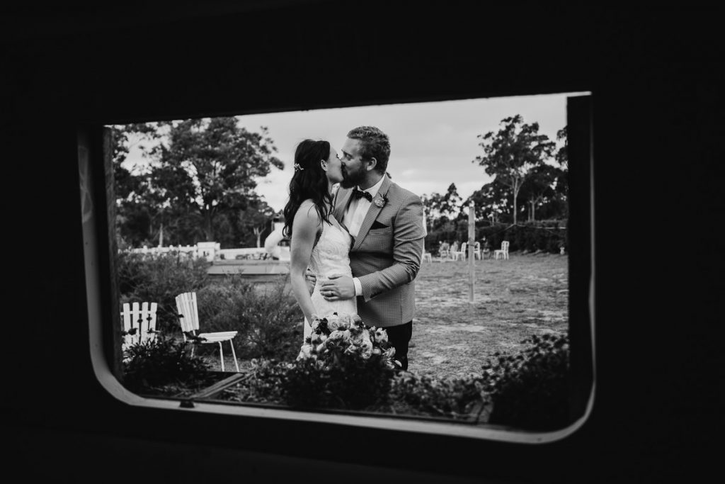 Black and white image of bride and groom kissing though the window of a shed at the Woods Farm Wedding Photographer South Coast