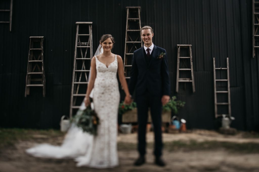 Couple pose in front of a blue shed with ladders hanging on the wall at their Woods Farm Wedding; South Coast Wedding Photographer