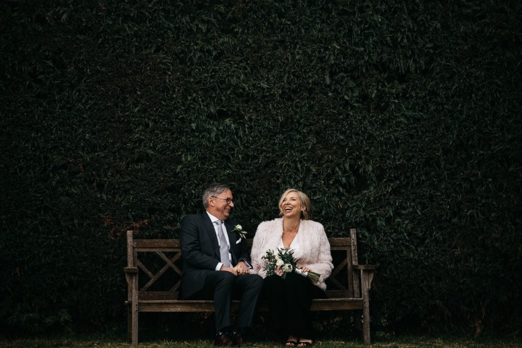 Mature bride and groom laughing while seated on a bench in front of a hedge at their wedding in Young