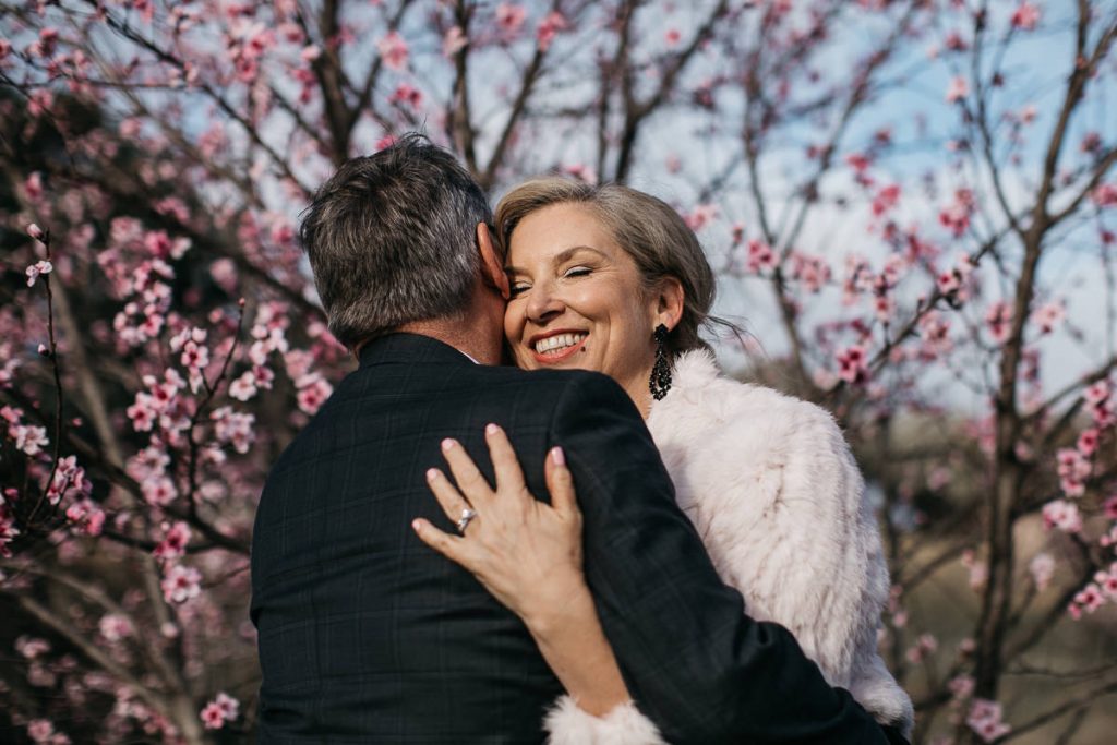 Mature couple hug under blossoming fruit trees at their Young Wedding; Country Wedding Photography